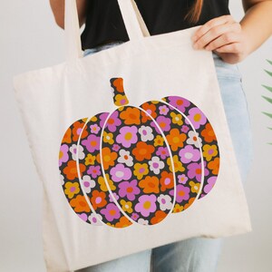 May include: A white canvas tote bag with a floral pumpkin design. The pumpkin is made up of pink, orange, and yellow flowers on a black background.