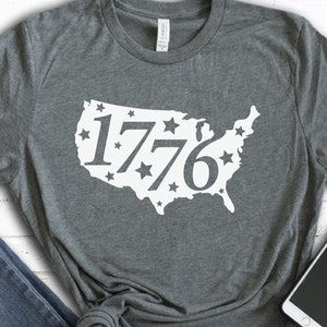 May include: Dark gray t-shirt featuring a white graphic of the United States map with the year "1776" inside. The map is outlined with stars. The shirt is made of a soft material.