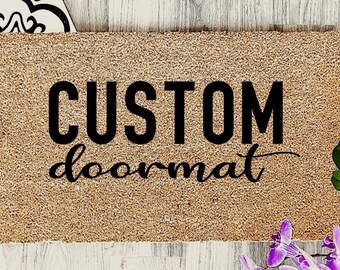 May include: A rectangular tan coir doormat featuring the black text CUSTOM in a bold sans-serif font and the word doormat in a black cursive script. The mat has a coarse, fibrous texture and is positioned on a light-colored wooden surface.