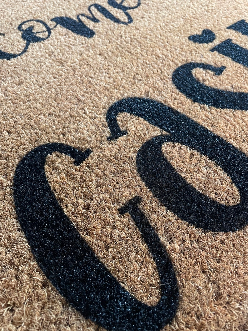 May include: Close-up of a brown coir doormat with the words "Welcome" in black lettering.
