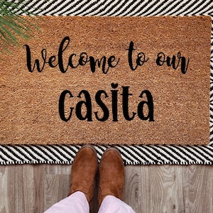 May include: Welcome to our casita doormat with black text on a brown coir background. The doormat is on a black and white striped rug with fringe.