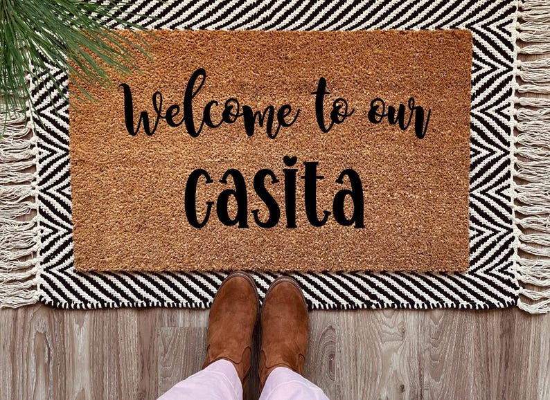 May include: Welcome to our casita doormat with black text on a brown coir background. The doormat is on a black and white striped rug with fringe.
