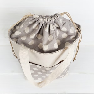 May include: A beige tote bag with a drawstring top and a gray and white polka dot pattern. The bag has two thick beige straps.