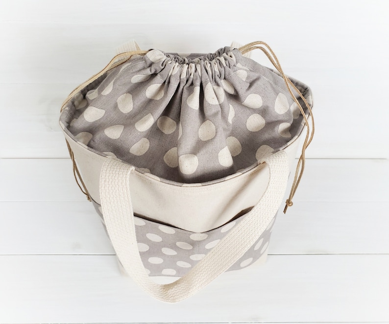 May include: A beige tote bag with a drawstring top and a gray and white polka dot pattern. The bag has two thick beige straps.
