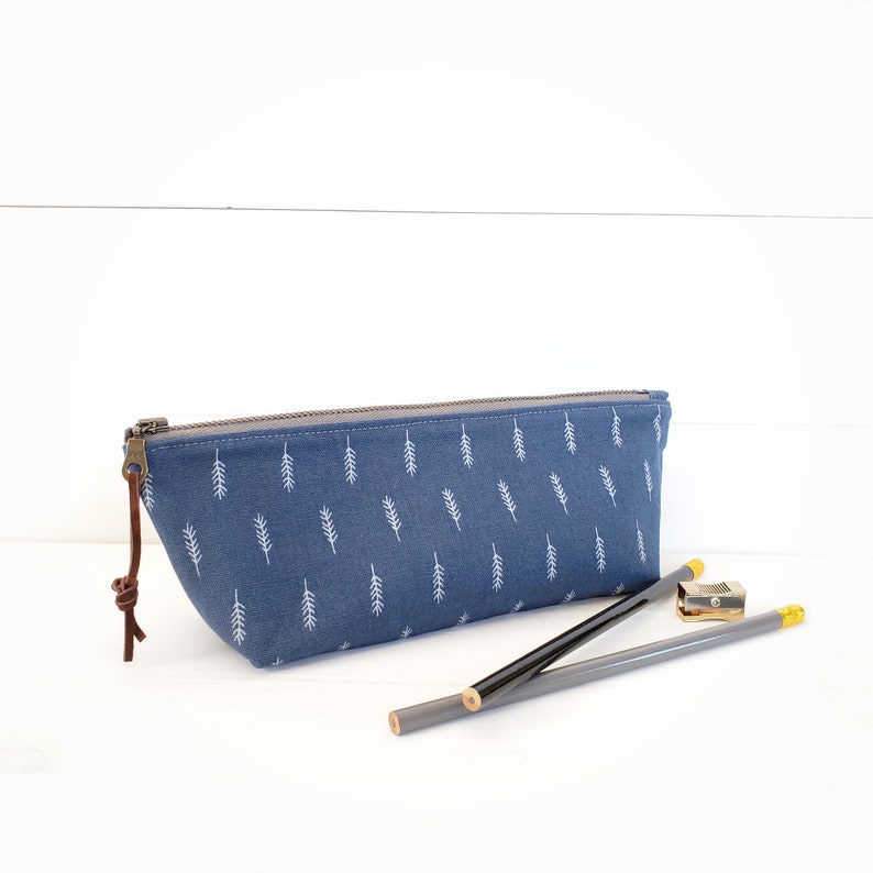 Op de afbeelding: Een blauwe potlood etui met een wit blad patroon en een bruine leren rits. Twee potloden en een puntenslijper liggen naast de etui.