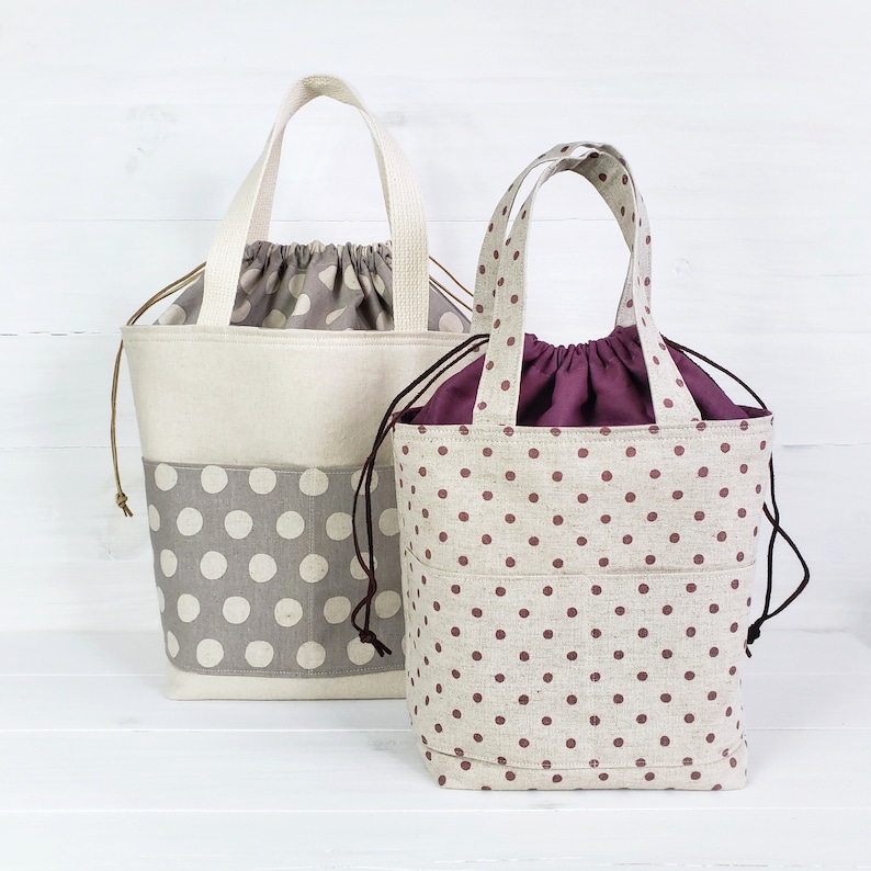May include: Two fabric tote bags with polka dot patterns. The larger bag is beige with gray polka dots and a brown drawstring. The smaller bag is beige with burgundy polka dots and a burgundy drawstring.