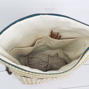May include: A yellow and white checkered zippered bag with a beige lining. The bag has multiple pockets and is open to reveal a ball of yarn and knitting needles.