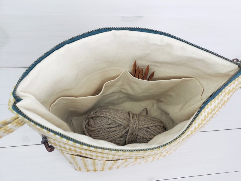 May include: A yellow and white checkered zippered bag with a beige lining. The bag has multiple pockets and is open to reveal a ball of yarn and knitting needles.