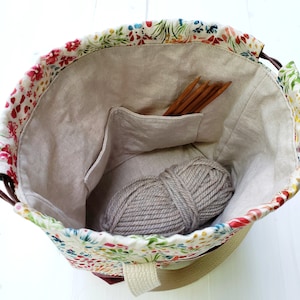 May include: A floral print drawstring bag with a beige lining. The bag is open and shows a ball of yarn and knitting needles inside. The bag has a beige cotton handle.