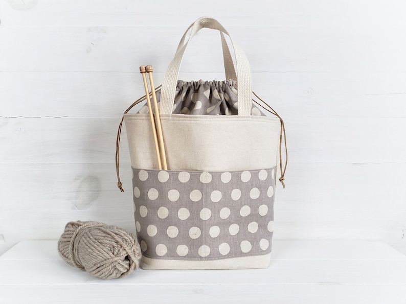 May include: A beige canvas tote bag with a gray and white polka dot fabric pocket. The bag has brown leather handles and a drawstring closure. The bag is filled with knitting needles and yarn.