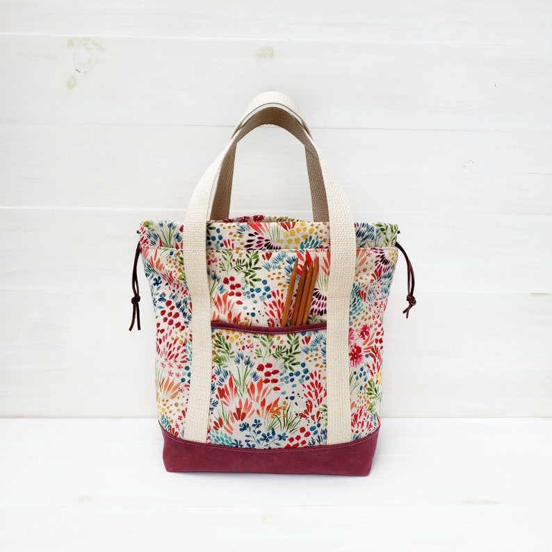 May include: A white tote bag with a floral print and a burgundy bottom. The bag has a large pocket on the front and is filled with knitting needles.