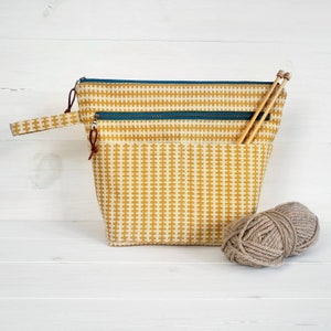 May include: A yellow and white checkered zippered pouch with a brown leather handle and two wooden knitting needles sticking out of the top. A ball of brown yarn is sitting in front of the pouch.