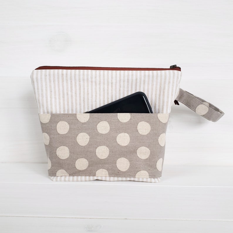 May include: A beige zippered pouch with a white and brown striped top and a bottom section with white polka dots on a beige background. A black cell phone is partially visible in the top pocket.