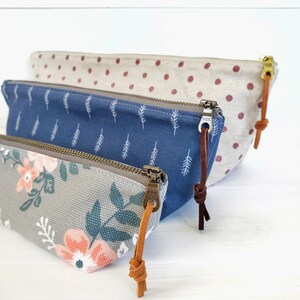 Op de afbeelding: Drie stoffen ritszakjes met verschillende patronen. Het bovenste zakje is beige met rode stippen, het middelste zakje is blauw met witte bladeren en het onderste zakje is grijs met een bloemenpatroon. Elk zakje heeft een bruine leren rits.