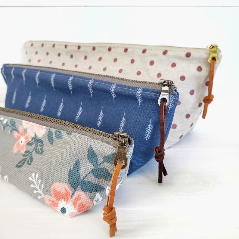 Op de afbeelding: Drie stoffen ritszakjes met verschillende patronen. Het bovenste zakje is beige met rode stippen, het middelste zakje is blauw met witte bladeren en het onderste zakje is grijs met een bloemenpatroon. Elk zakje heeft een bruine leren rits.