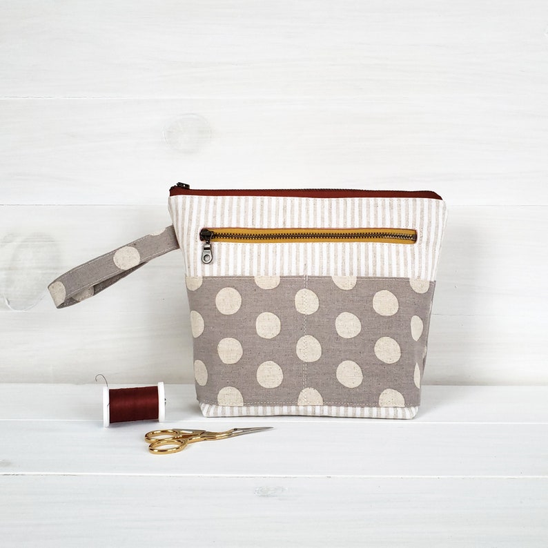 May include: A gray and white polka dot zippered pouch with a brown zipper pull and a brown and white striped top. The pouch has a handle and is sitting on a white wooden surface.