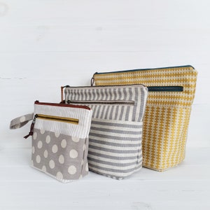 May include: Three fabric zippered pouches in different sizes and patterns. The largest pouch is yellow with a white geometric pattern, the middle pouch is gray with white stripes, and the smallest pouch is gray with white polka dots.