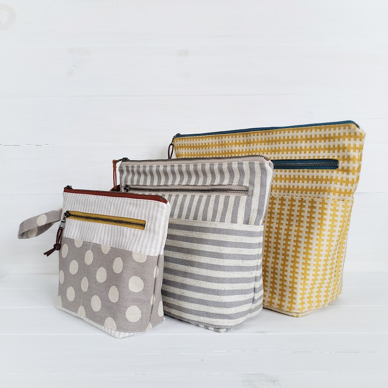 May include: Three fabric zippered pouches in different sizes and patterns. The largest pouch is yellow with a white geometric pattern, the middle pouch is gray with white stripes, and the smallest pouch is gray with white polka dots.