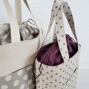 May include: Two tote bags with brown polka dots on a natural linen fabric. The larger bag has a cream colored lining and the smaller bag has a burgundy lining.