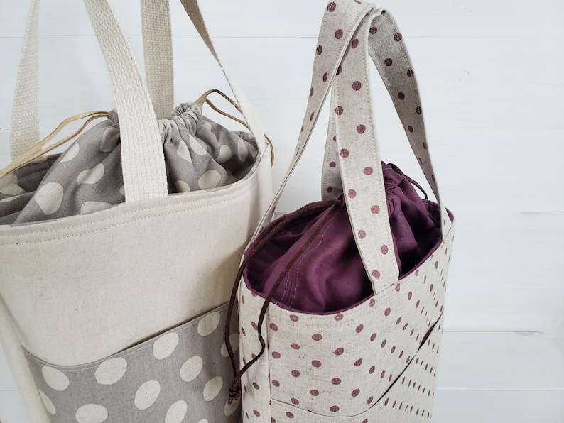 May include: Two tote bags with brown polka dots on a natural linen fabric. The larger bag has a cream colored lining and the smaller bag has a burgundy lining.