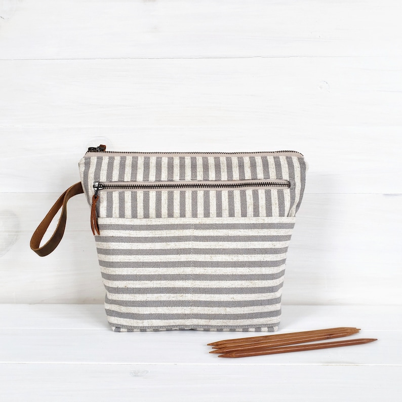 May include: A gray and white striped zippered pouch with a brown leather handle. The pouch has two zippered compartments and a large pocket on the front. The pouch is sitting on a white wooden surface with four wooden knitting needles in front of it.