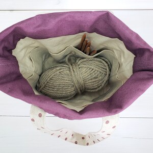 May include: A purple tote bag with a white polka dot lining and handles. The bag is open and contains a ball of beige yarn and wooden knitting needles.