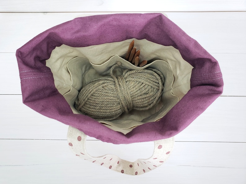 May include: A purple tote bag with a white polka dot lining and handles. The bag is open and contains a ball of beige yarn and wooden knitting needles.