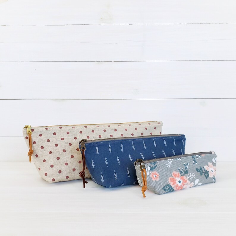 Op de afbeelding: Drie stoffen ritszakjes in verschillende maten en patronen. Het grootste zakje is rood met witte stippen, het middelste zakje is blauw met een wit bladpatroon en het kleinste zakje is grijs met een roze en groen bloemenpatroon.