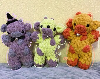 May include: Three handmade crochet plush toys featuring soft yarn textures. The left toy is purple with a black witch hat and bell. The center toy is light green with purple spots and a bell. The right toy is golden brown with orange spots and a bell.
