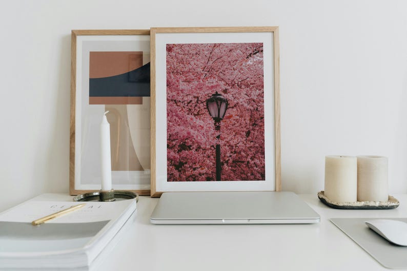 Puede incluir: Una impresi&oacute;n enmarcada de un cerezo en flor rosa con una farola en el centro. La impresi&oacute;n est&aacute; sobre un escritorio blanco con un port&aacute;til, un cuaderno, un portavelas y dos velas.