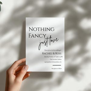 May include: A white wedding invitation with the text "NOTHING FANCY just love" in black script. The invitation includes details for the wedding of Rachel & Ross, including the date, time, and location. The invitation is held by a hand.