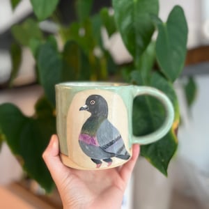 Op de afbeelding: Een lichtgroene keramische mok met een handgeschilderde illustratie van een grijze duif met een paarse vlek op de nek. De duif is in profiel afgebeeld tegen een crèmekleurige ovale achtergrond. De mok heeft een stevig handvat en een gladde afwerking.