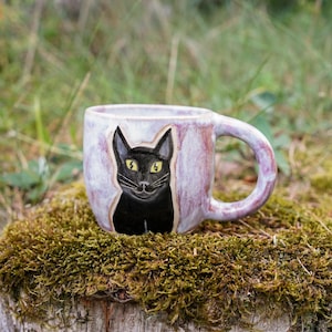 May include: A ceramic mug with a light purple and white mottled glaze featuring a hand-painted illustration of a black cat with bright yellow eyes on the front. The mug has a rounded body and a curved handle extending from the right side.