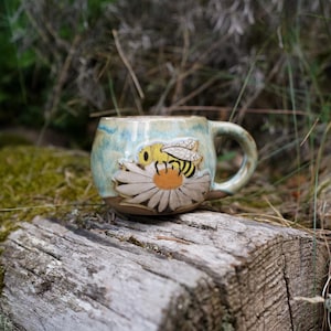 May include: A ceramic mug with a light blue and tan glaze featuring a raised, hand-painted design of a yellow and black bee resting on a white daisy with a yellow center. The mug has a rounded body and a curved handle on the right side.