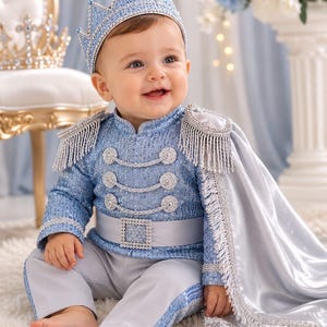 Könnte beinhalten: Eine hellblaue Paillettenjacke mit silbernem Flechtbesatz, Schulterklappen und einem passenden Gürtel mit quadratischer Strassschnalle. Das Outfit umfasst eine hellgraue Hose und einen langen, silbernen Satinumhang mit Fransenbesatz sowie eine Krone.