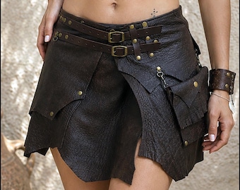 Könnte beinhalten: Ein dunkelbrauner Rock aus strukturiertem Leder mit asymmetrischem Saum, zwei horizontalen Schnallenriemen an der Taille mit Messingbeschlägen und einer kleinen abnehmbaren Tasche, die seitlich mit einer Metallkette und Druckknöpfen befestigt ist.