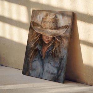 May include: Canvas art print of a person wearing a cowboy hat and denim shirt. The artwork features a neutral color palette with a focus on the subject's face and long hair. The hat is a light brown color, and the shirt is a faded blue.