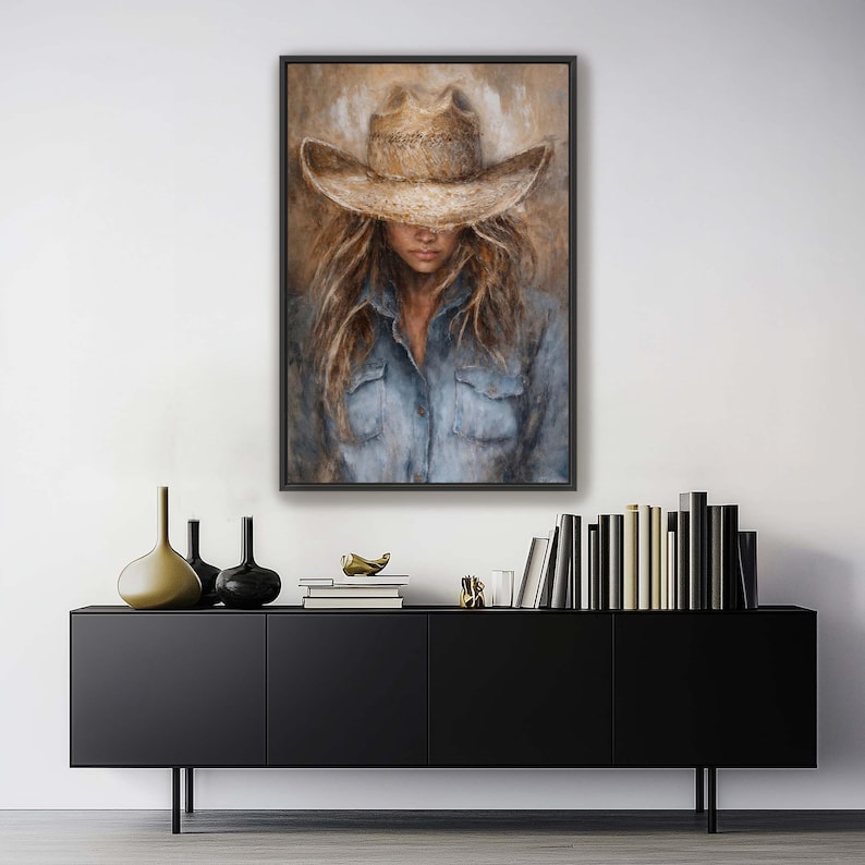 May include: A framed painting depicts a person wearing a cowboy hat and denim shirt. The artwork features a neutral color palette with shades of brown, beige, and blue. The subject's face is partially obscured by the hat, creating a sense of mystery.