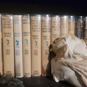 May include: A shelf of vintage Nancy Drew mystery books with off-white covers and blue illustrations. Titles include "The Broken Locket" and "The Ivory Charm." A white angel statue rests on the shelf.