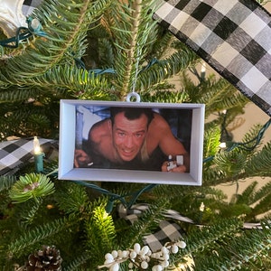 May include: A white picture frame ornament with a black and white checkered ribbon hanging on a green Christmas tree. The picture frame contains a photo of a man with a surprised expression.
