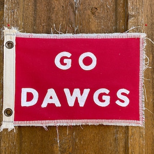 May include: A red fabric flag with white lettering that says "GO DAWGS" hanging on a wooden surface.