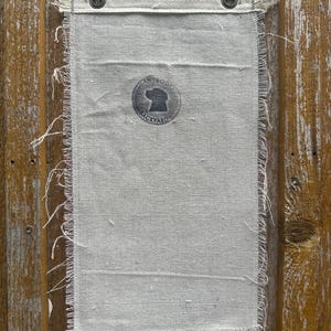 Op de afbeelding: Een rechthoekige, cr&egrave;mekleurige canvas banner met gerafelde randen en een versterkte zoom. De banner heeft een cirkelvormig logo met een honden silhouet en de tekst "ANTIQUE BACKYARD". Twee metalen ringen zijn in de bovenzoom geplaatst.