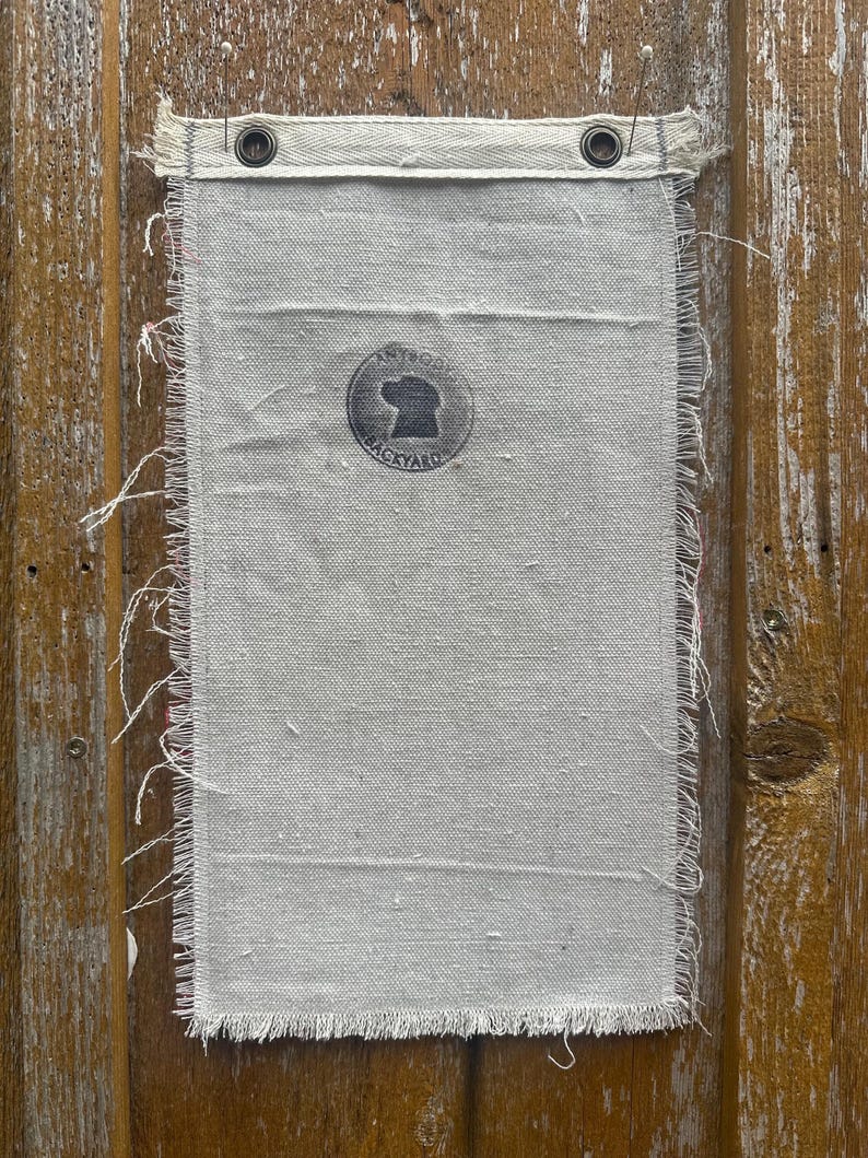Op de afbeelding: Een rechthoekige, cr&egrave;mekleurige canvas banner met gerafelde randen en een versterkte zoom. De banner heeft een cirkelvormig logo met een honden silhouet en de tekst "ANTIQUE BACKYARD". Twee metalen ringen zijn in de bovenzoom geplaatst.