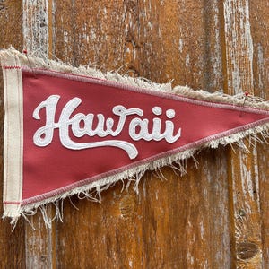 May include: A triangular pennant in a muted red color featuring the word Hawaii in white script lettering. The pennant has a cream-colored canvas header strip on the left side and frayed fabric edges along the top and bottom borders.