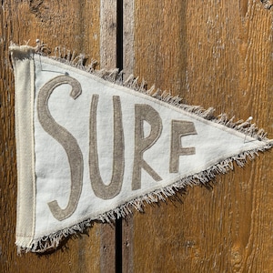May include: A white pennant with frayed edges and the word "SURF" in brown embroidered letters. The pennant is pinned to a wooden surface.