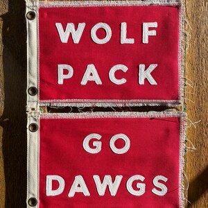 Op de afbeelding: Twee rode stoffen vlaggen met witte tekst. De bovenste vlag leest "WOLF PACK" en de onderste vlag leest "GO DAWGS". De vlaggen hebben een witte rand met ogen en zijn vastgespeld op een houten oppervlak.
