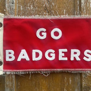 Op de afbeelding: Een rode vlag met de witte woorden "GO BADGERS". De vlag heeft een gerafelde rand en oogjes aan de linkerkant. De vlag is rechthoekig en lijkt van stof te zijn.
