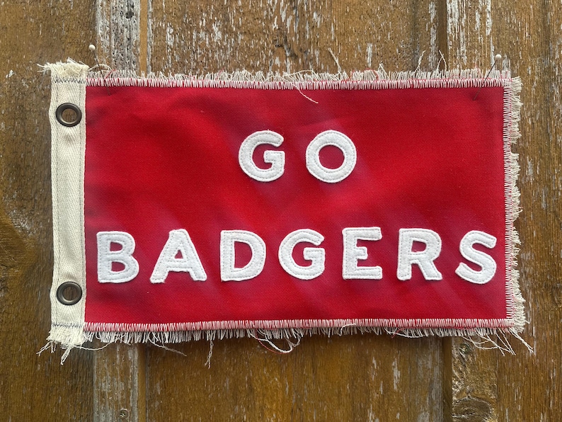 Op de afbeelding: Een rode vlag met de witte woorden "GO BADGERS". De vlag heeft een gerafelde rand en oogjes aan de linkerkant. De vlag is rechthoekig en lijkt van stof te zijn.