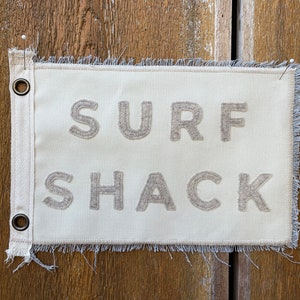 May include: A white canvas sign with frayed edges and metal eyelets. The sign reads "SURF SHACK" in beige embroidered lettering.