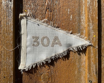 May include: A triangular, off-white fabric pennant with the number "30A" stitched in a light brown thread. The pennant has a frayed edge and is pinned to a weathered, wooden surface with a yellow pin.